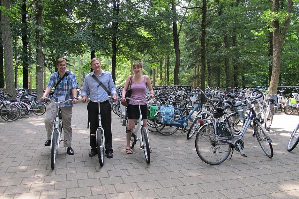 3 people riding bicycles in a park with many other parked bicycles